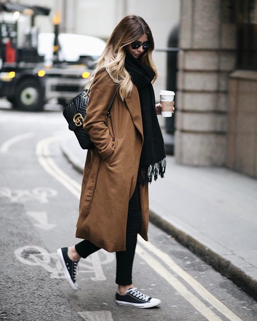 Emma Hill wears black and white converse with black jeans, an oversized black frayed-end scarf, and a cosy brown overcoat. This style is finished off with a leather handbag and shades. Brands not specified.