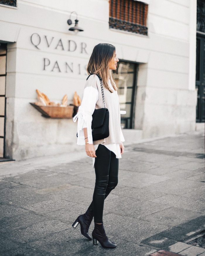 Alba Hervas wears tight leather leggings with sleek black boots, a layered white blouse with cutaway sleeves, and a beige top. Alba pairs this look with a lush black handbag. Brands not specified.